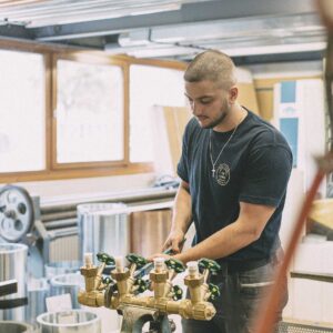 Ein junger Mann mit kurzem Haar arbeitet konzentriert an einem Rohrventil in einer Werkstattumgebung, trägt ein dunkelblaues T-Shirt und eine Halskette